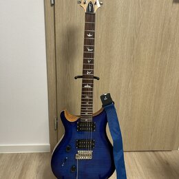 Modrá elektrická kytara s dumpedým vrchem v přírodní barvě dřeva, umístěná na černém stojanu. Na hlavě je bílé logo "Cort" a nápis "Custom". Hmatník…