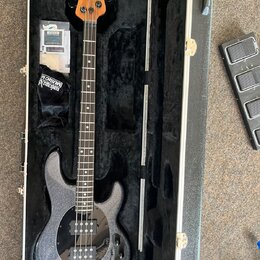 Stříbrná elektrická basová kytara s třpytivým povrchem v černém tvrdém kufru. Na hlavě hmatníku je čitelné logo Music Man. Vedle krku nástroje leží č…