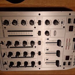 Stříbrný DJ mixér s nápisy BEHRINGER a VMX 100. Na horní části jsou ovládací prvky pro MAIN OUT, OUT A a OUT B, vpravo je vidět logo XPO. Přístroj ob…