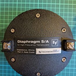 Černý kulatý komponent s texturovaným povrchem, připevněný čtyřmi šrouby. Uprostřed je umístěn štítek s nápisy „Diaphragm S/A“, „for High-Frequency T…