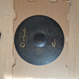 Kruhový činely z tmavého kovaného kovu s viditelnou strukturou kladivování. Na povrchu je žlutý nápis „Turkish“ a pod ním menší text „Handmade cymbal…