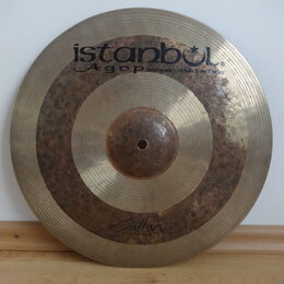 Kovový činel zlatavé barvy s viditelným Oxidováním a tmavými skvrnami v prostřední části. V horní části je černý nápis istanbul Agop Handmade cymbals…