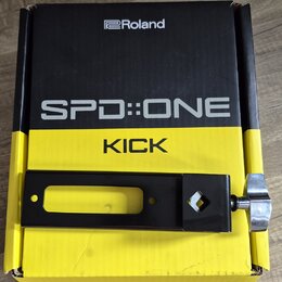 Černý kovový držák s utahovacím šroubkem z chromovaného kovu, položený na žlutočerné krabici. Na krabici jsou bílé a černé nápisy „Roland“, „SPD::ONE…
