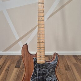 Elektrická kytara s hnědým dřevěným korpusem a světle hnědým javorovým hmatníkem. Na hlavě je viditelné logo Fender. Kytara má černý mramorovaný pick…