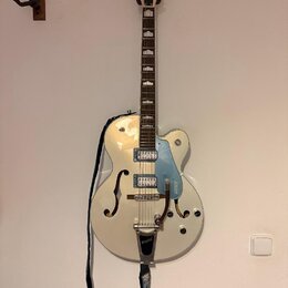 Bílá polodupatá elektrická kytara s modrým pickguardem a chromovaným hardwarem. Na hlavě hmatníku je nápis GRETSCH a na pickguardu je svisle umístěný…