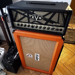 Černý kytarový zesilovač s logem EVH a označením 5150 III TUBE EL34. Pod ním stojí oranžový kytarový kombozesilovač s logem ORANGE. Oba přístroje jso…
