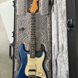 Modrá elektrická kytara typu Stratocaster s bílým pickguardem a javorovým hmatníkem v otevřeném černém tvrdém kufru s šedým polstrováním. Na hlavě ná…
