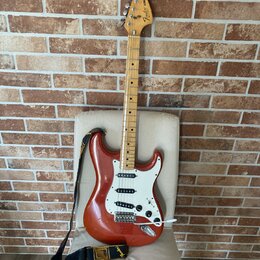 Elektrická kytara typu Stratocaster v červeně hnědé barvě s bílým pickguardem a světle hnědým hnedobým hmatníkem. Na hlavě je viditelné logo Fender S…