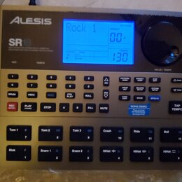 Elektronický bicí automat značky Alesis, model SR18. Zařízení má stříbrno-šedý korpus s černým ovládacím panelem a modrým podsvíceným displejem, na k…