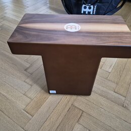 Hnědý dřevěný cajón s logem MEINL v bílém kruhu na horní hraně. Nástroj má kompaktní tvar s vyčnívající horní deskou a užším spodním tělem. Povrch dř…