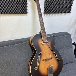 Kytara typu archtop s tmavě hnědým sunburst povrchem a bílým lemováním. Na hlavě jsou vidět čtyři ladičky a dva červené dekorativní prvky. Tělo má dv…