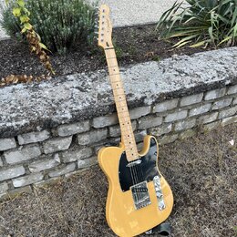Elektrická kytara typu Telecaster v žlutém provedení s černým pickguardem. Na hlavě hmatníku je viditelné logo Squier. Nástroj vykazuje známky použív…