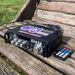 Černý kompaktní audio zesilovač s kovovým povrchem a ovládacími knofíky. Na horní části je nápis 300W+300W a náleпка s fialovými postavami. Na přední…