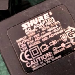 Černý síťový adaptér s bílým textem. Je na něm uveden nápis SHURE® AC ADAPTER, MODEL NO: PS31E, INPUT: 230V ~ 50Hz 0.06A a OUTPUT: 15V = 400mA. Dále…