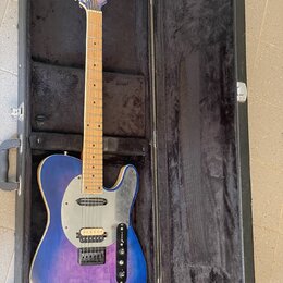 Modrá elektrická kytara typu Telecaster s javorovým hmatníkem v černém tvrdém kufru. Na korbisku je viditelné logo "Fender". Kytara má stříbrný plech…