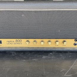 Kytarový zesilovač v černém obalu s šedou textilní mřížkou. Zlatý přední panel nese nápis „replica 800 LEAD SERIES“. Na panelu jsou umístěny dva vypí…