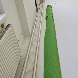 Na fotografii je stříbrný hliníkový mřížový truss v poloze vleže na podlaze. Vedle něj leží dlouhý zelený textilní kryt nebo přepravní vak. Na vybave…