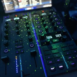 Černý DJ kontrolér s několika knoflíky, posuvníky a podsvícenými tlačítky. Na přístroji je svisle napsán bílý text TRAKTOR Z1. V pozadí je vidět zapn…