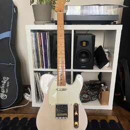 Elektrická kytara typu Telecaster v krémové barvě s bílým pickguardem a světle hnědým hnedem hmatníkem. Na hlavě je viditelné logo Squier. Nástroj st…