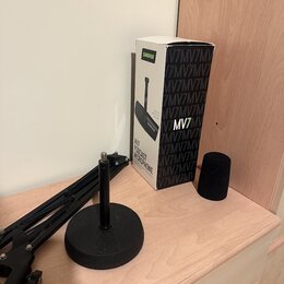 Na fotografii je krabice od mikrofonu s nápisy SHURE, MV7 a MV7 PODCAST MICROPHONE. Vedle krabice je černý pěnový popfilter a černý mikrofonní ramení…