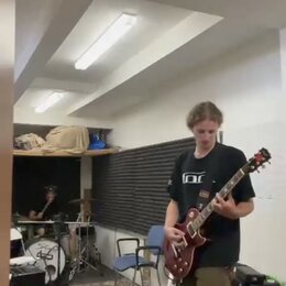 Muž v černé košili a khaki šortkách hraje na červenou elektrickou kytaru s tvarem singlecut. Na hlavě kytary je viditelné bílé logo, které je však ne…
