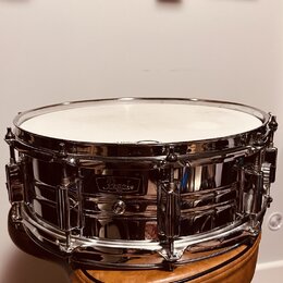 Stříbrný kovový malý buben s bílým koženem, umístěný na hnědém polstrovaném sedadle. Na korpusu je viditelné logo "Pearl". Povrch nástroje je vysoce…