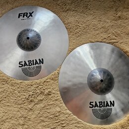 Dvě stříbrné činely značky Sabian s matným povrchem. Levý činel nese nápisy FRX, Hats 14"/35 cm a logo SABIAN. Pravý činel má pouze logo SABIAN. Oba…