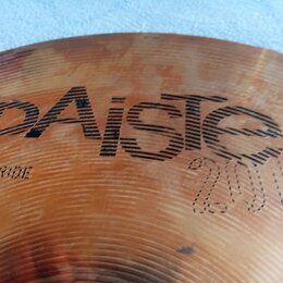 Detailní záběr na povrch činchy v bronzové barvě. Na povrchu je vidět nápis „paiste“ a pod ním označení „20" RIDE“. V pravé dolní části je vyryto „20…