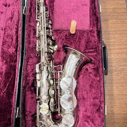 Stříbrný saxofon v černém kufru s fialovým polstrováním. Nástroj vykazuje známky opotřebení v podobě drobných škrábanců a znečištění povrchu. Na těle…