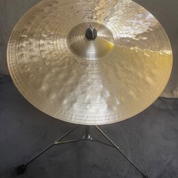 Zlatý kovový činel umístěný na černém stativu. Na povrchu je nápis „Paiste“ a označení „Dry Ride 20"“. Povrch činelu je zdobný koncentrickými rýhami…