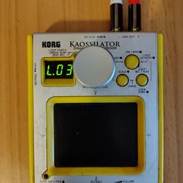 Stříbro-žlutý přenosný syntetizátor s nápisy KORG a KAOSSILATOR DYNAMIC SYNTHESIZER. Zařízení má malý digitální displej s nápisem L.03, jeden velký s…