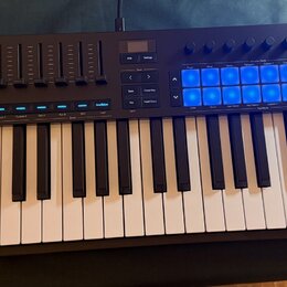 Černý MIDI kontrolér s klaviaturní klávesnicí. Na levé straně je bílý nápis LAUNCHKEY a řada posuvníků. V pravé horní části se nachází mřížka 16 modř…