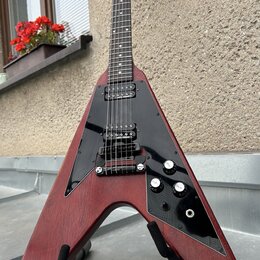 Elektrická kytara s Flying V tvarem těla v červeném dřevěném provedení a černým pickguardem. Na hmatníku jsou dva humbuckery, tři ovládací knofíky a…