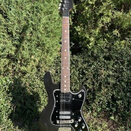 Černá elektrická kytara s černým pickguardem a hnědým hmatníkem. Na hlavě krku je viditelné bílé logo Fender. Nástroj má dva snímače, tři ovládací kn…