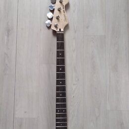 Fotografie zachycuje hmatník a hlavu basové kytary z přírodního dřeva s tmavým hmatníkem. Na hlavě je vidět nápis Squier JAZZ BASS a čtyři kovové lad…
