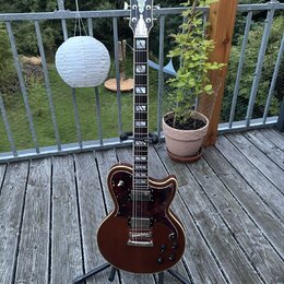 Tmavně hnědá elektrická kytara s tvarem single-cut, umístěná na černém stojanu. Na hlavě je bílé logo D'Angelico, korpus má tmavě červený perleťový p…
