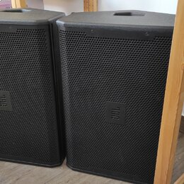 Dva černé reproduktory v plastových skříních s kovovými mřížkami. Na přední straně každého reproduktoru je umístěn čtvercový štítek s nápisem JBL. Po…