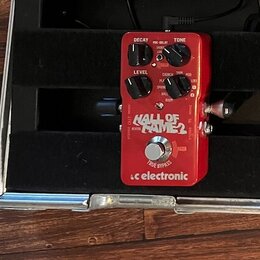 Červený efekt pedál s nápisy HALL OF FAME 2 a tc electronic. Na horní části jsou čtyři ovládací knoflíky s popisky DECAY, PRE-DELAY, TONE a LEVEL. V…
