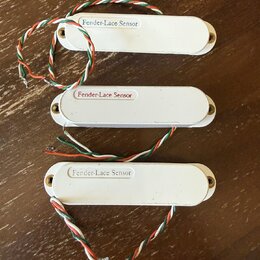 Tři bílé elektrické snímače s oválným tvarem a montážními šrouby po stranách. Každý snímač má na horní části nápis Fender-Lace Sensor. Ze snímačů ved…
