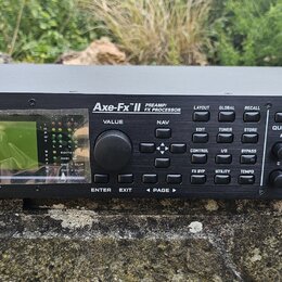 Černý rackový procesor s nápisy FRACTAL AUDIO SYSTEMS a Axe-Fx II PREAMP/FX PROCESSOR. Zařízení má displej, několik tlačítek s popisky jako LAYOUT, G…