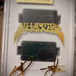 Dva černé humbuckery v bílém plastovém obalu, který je zabalen v průhledné fólii. Na horním okrape obalu je žlutý štítek s nápisem FISHMAN Signature…