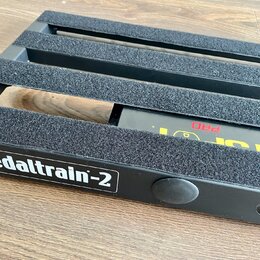 Černý kovový pedalboard s nápisem pedaltrain-2 v bílém rámečku. Horní lišty jsou potaženy šedým protiskluzovým materiálem. Pod deskou je vidět část č…