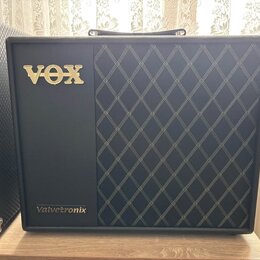 Černý kytarový kombozesilovač s pravou stranou pokrytou černou tkaninou s bílým kosočtvercovým vzorem. V horní levé části je umístěno zlaté logo VOX…