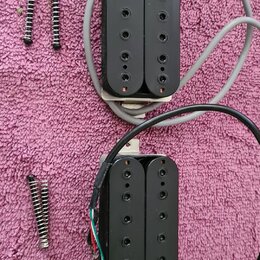 Dvě černé humbucker snímače s kovovými základnami a připojenými kabely v šedé a černé barvě. Vedle snímačů jsou položeny čtyři montážní pružiny s šro…
