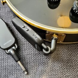 Dvě černé bezdrátové vysílače/přijímače s kovovými jack konektory. Na levém zařízení je nápis RECEIVER, na pravém jsou nápisy POWER PAIR, CHARGE ID a…