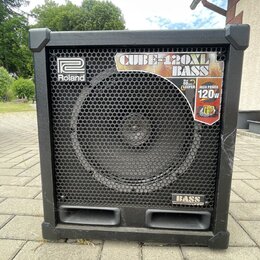 Černý basový kombozesilovač s kovovou ochrannou mřížkou přes reproduktor. Na přední straně jsou viditelná loga a nápisy: "Roland", "CUBE-120XL BASS",…