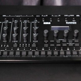 Černý hybridní bicí modul s nápisy KORG a drumlogue HYBRID DRUM MACHINE. Zařízení obsahuje několik otočných potenciometrů, digitální displej a řadu o…