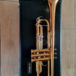Zlatý mosazný trumpet položený na černém kufru. Na kufru je bílý nápis YAMAHA v obdélníkovém rámečku. Nástroj vykazuje známky používání v podobě drob…