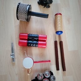 Sestava různých perkusních nástrojů na světlé podlaze. Soubor obsahuje černý buben s kovovými plátky, červené shakerové trubice, dřevěný guiro s modr…