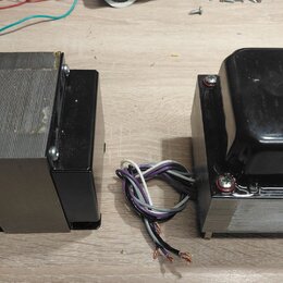 Dvě elektrické transformátory s kovovými jádry a černým krytím. Levý kus má část jádra odkrytou, pravý je z horní strany zakryt černým plastovým nebo…